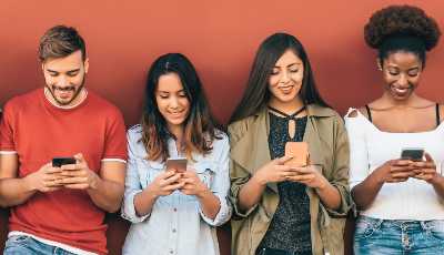 Four young people leaning against a wall and smiling while looking at their phones
