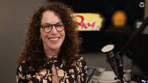 Shelli Sonstein smiling sitting in front of microphone in radio station with Q104 logo in background.