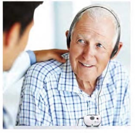Image of the Mino Personal Amplifier, a small, handheld black device with clearly visible buttons and controls, and a cord leading to over-the-ear headphones worn by a person.